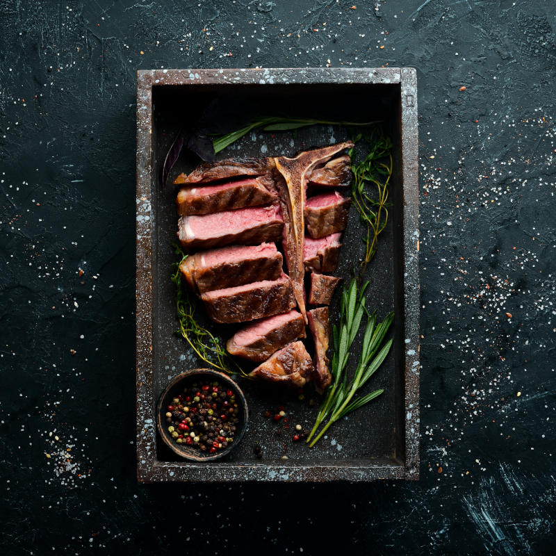 T-Bone Steak auf einer Schieferplatte umringt von Kräutern und einem Schüsselchen mit buntem Pfeffer